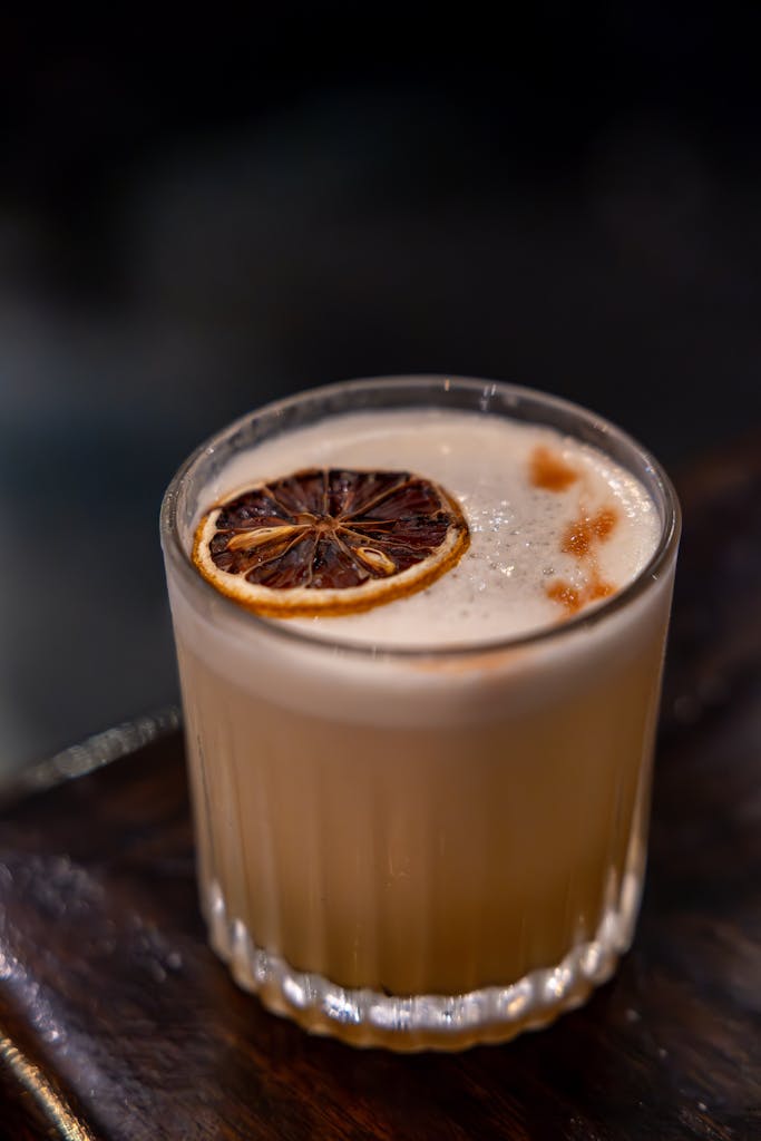 Close-up of a whiskey sour cocktail with a slice of dried citrus in a glass tumbler on a wooden surface.