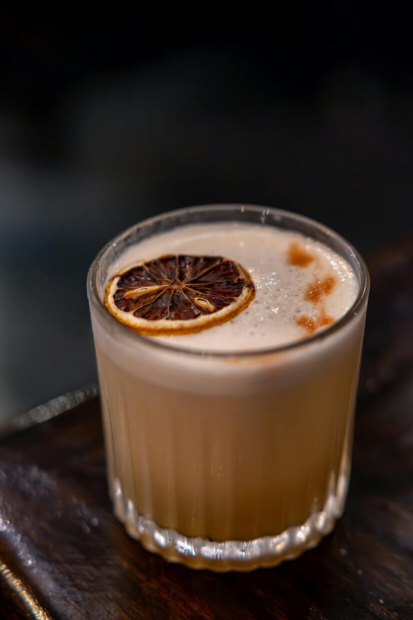 Close-up of a whiskey sour cocktail with a slice of dried citrus in a glass tumbler on a wooden surface.