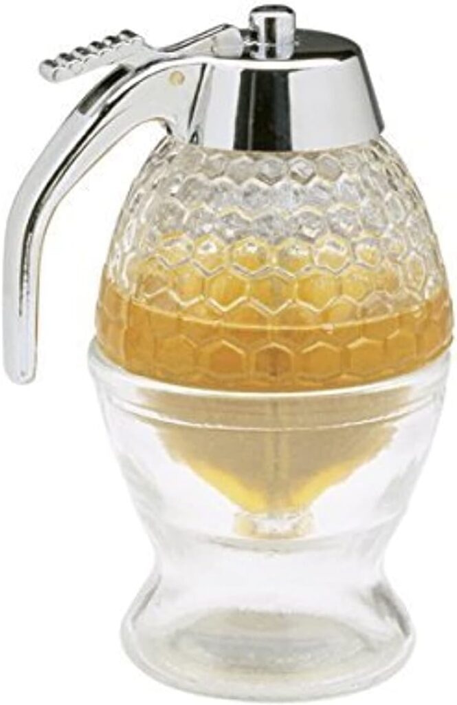 Glass maple syrup dispenser with a honeycomb-patterned top and chrome-plated handle and lid. The clear container shows golden syrup inside and sits atop a matching glass base designed to catch drips.