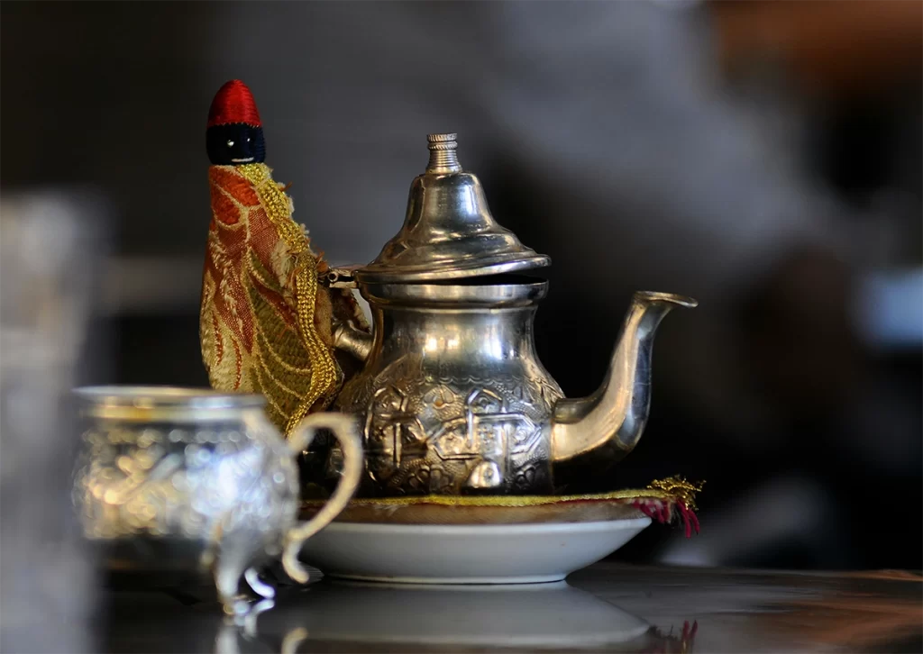 An ornate silver teapot with traditional engravings rests on a white saucer beside a matching tea cup, with a decorative fabric cozy and a red-topped stopper adding cultural flair. This inviting tea setup reflects a local tea experience, ideal for seniors seeking comforting and authentic tea choices while travelling.
