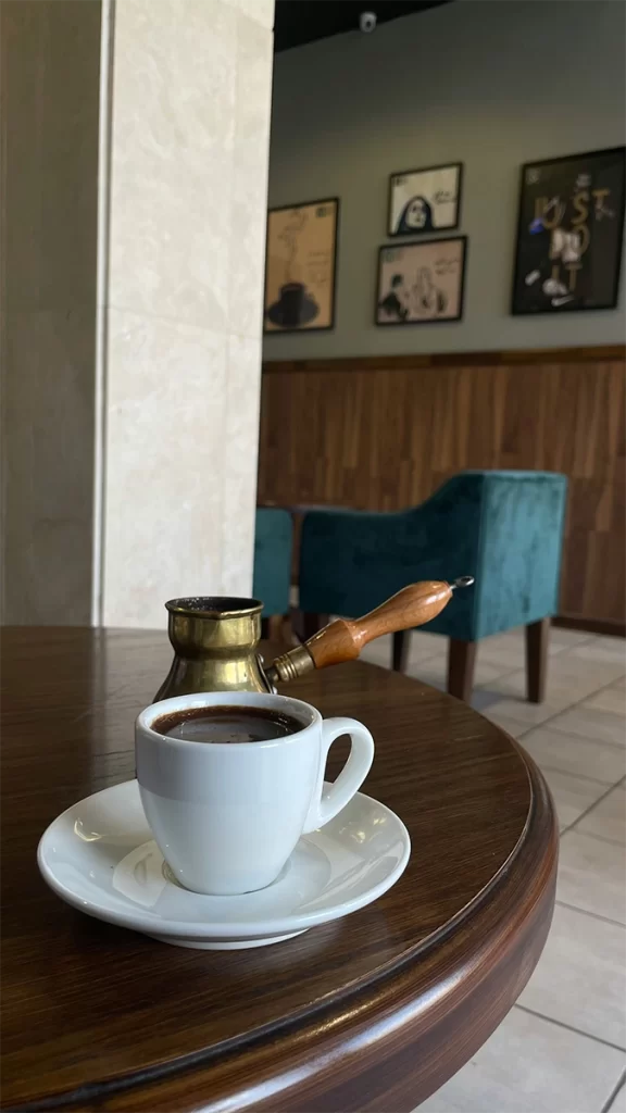 A cup of rich, dark Turkish coffee sits on a white saucer beside a traditional brass cezve with a wooden handle, set on a polished wooden table in a cozy café. This simple, single-origin brew is a safe coffee option for seniors, offering bold flavor without added sugar or dairy
