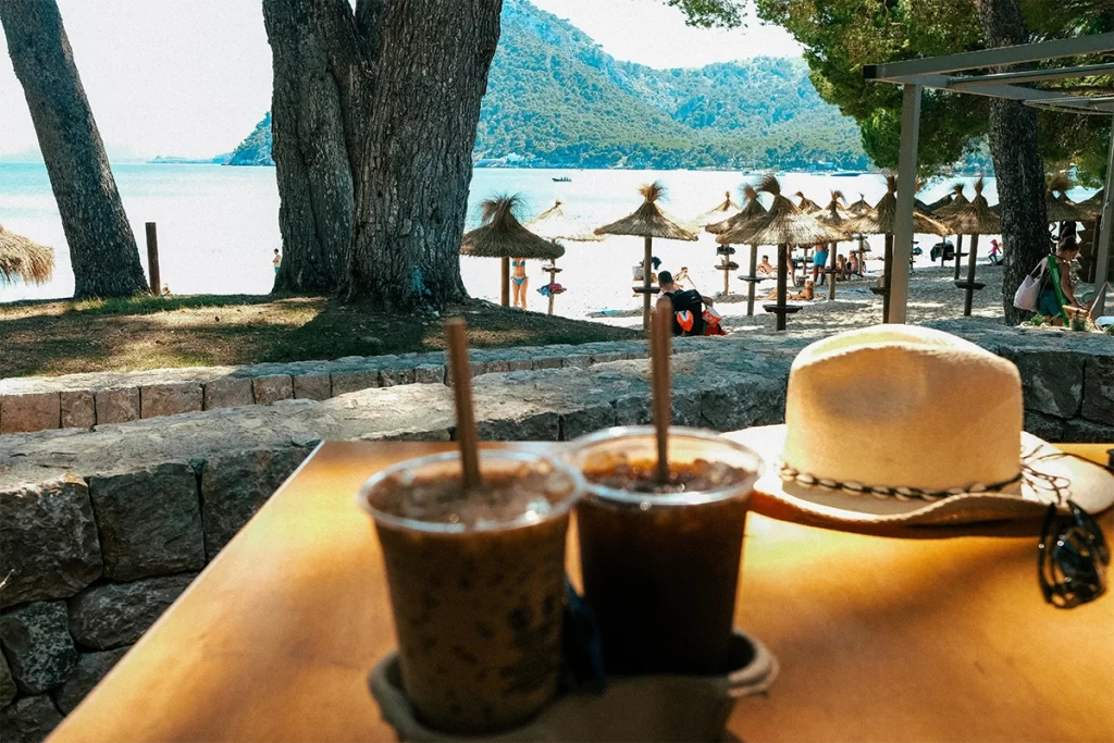 Two iced coffees sit on a wooden table next to a white sunhat and sunglasses, overlooking a serene beach with straw umbrellas, pine trees, and distant mountains. The relaxed seaside café setting suggests a comfortable, senior-friendly destination where ordering coffee abroad can be enjoyed with ease.