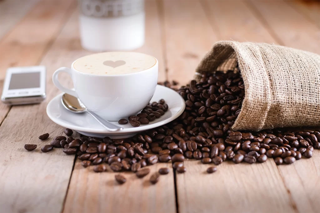 A frothy cup of coffee with a heart design in the foam sits on a saucer surrounded by roasted coffee beans spilling from a burlap sack on a wooden table. This cozy setup reflects the growing interest in healthy coffee sweeteners in Canada, where natural options like maple syrup offer a flavorful and less processed alternative to refined sugar.