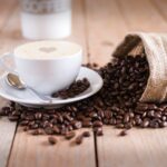 A white cup of coffee sits beside roasted coffee beans spilling from a burlap sack across a wooden table. The photo provides a general coffee visual for a guide on how to read a coffee bag label but no label details are visible.