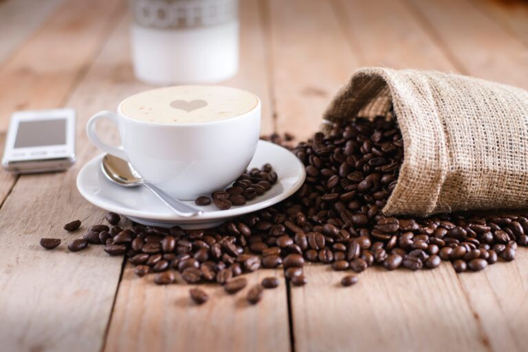 A white cup of coffee sits beside roasted coffee beans spilling from a burlap sack across a wooden table. The photo provides a general coffee visual for a guide on how to read a coffee bag label but no label details are visible.