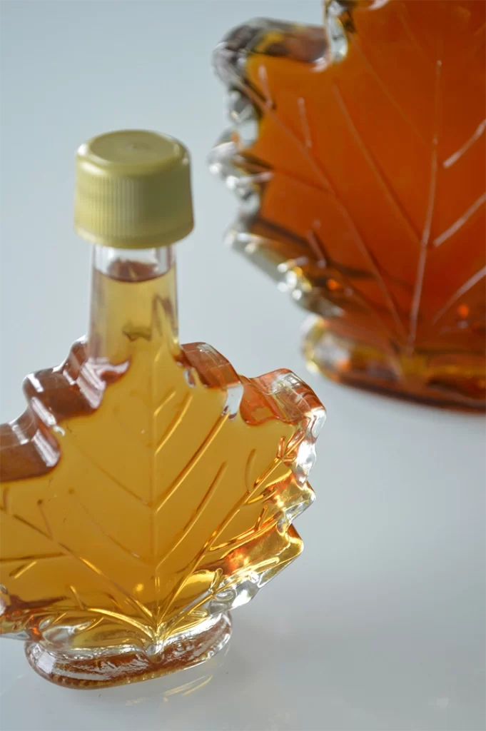Clear maple leaf-shaped glass bottles filled with amber-colored syrup, often used as a natural sweetener and a popular alternative to processed sugar in healthy coffee options for older adults. One bottle is in sharp focus while the other is slightly blurred in the background.