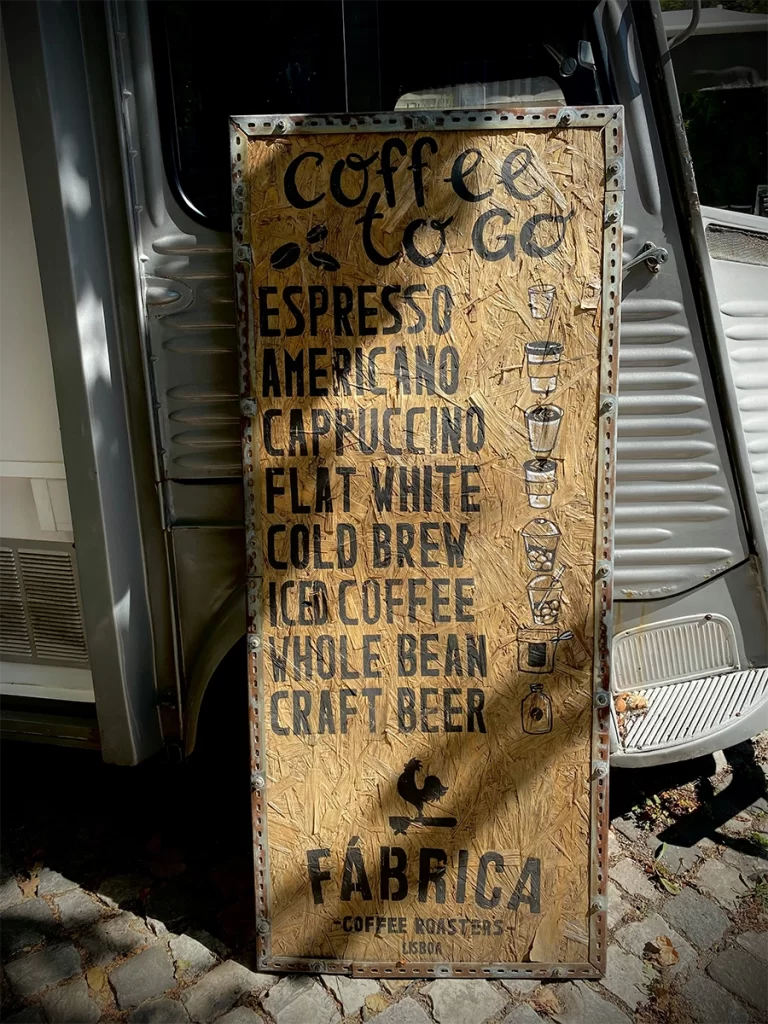A rustic wooden sign outside a coffee van lists drink options including espresso, americano, cappuccino, flat white, cold brew, iced coffee, whole bean, and craft beer, with playful hand-drawn icons beside each item. This clear and visual menu from Fábrica Coffee Roasters in Lisbon makes ordering coffee abroad more approachable for seniors. this is ow to order coffee abroad for seniors
