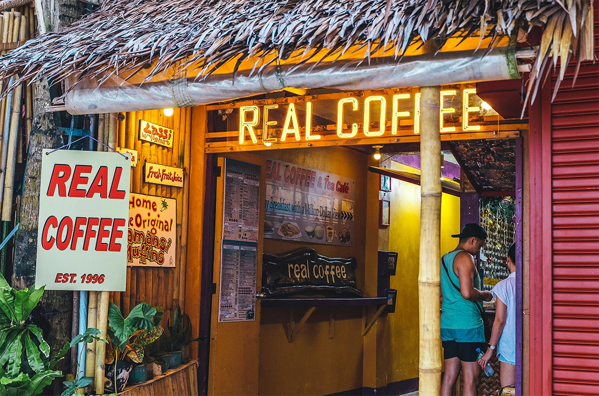 A vibrant tropical café with a thatched roof and glowing "REAL COFFEE" sign welcomes visitors with its bold signage and colorful menu board. The casual setup hints at locally made drinks and potentially healthy coffee options abroad with fresh ingredients and fewer additives.