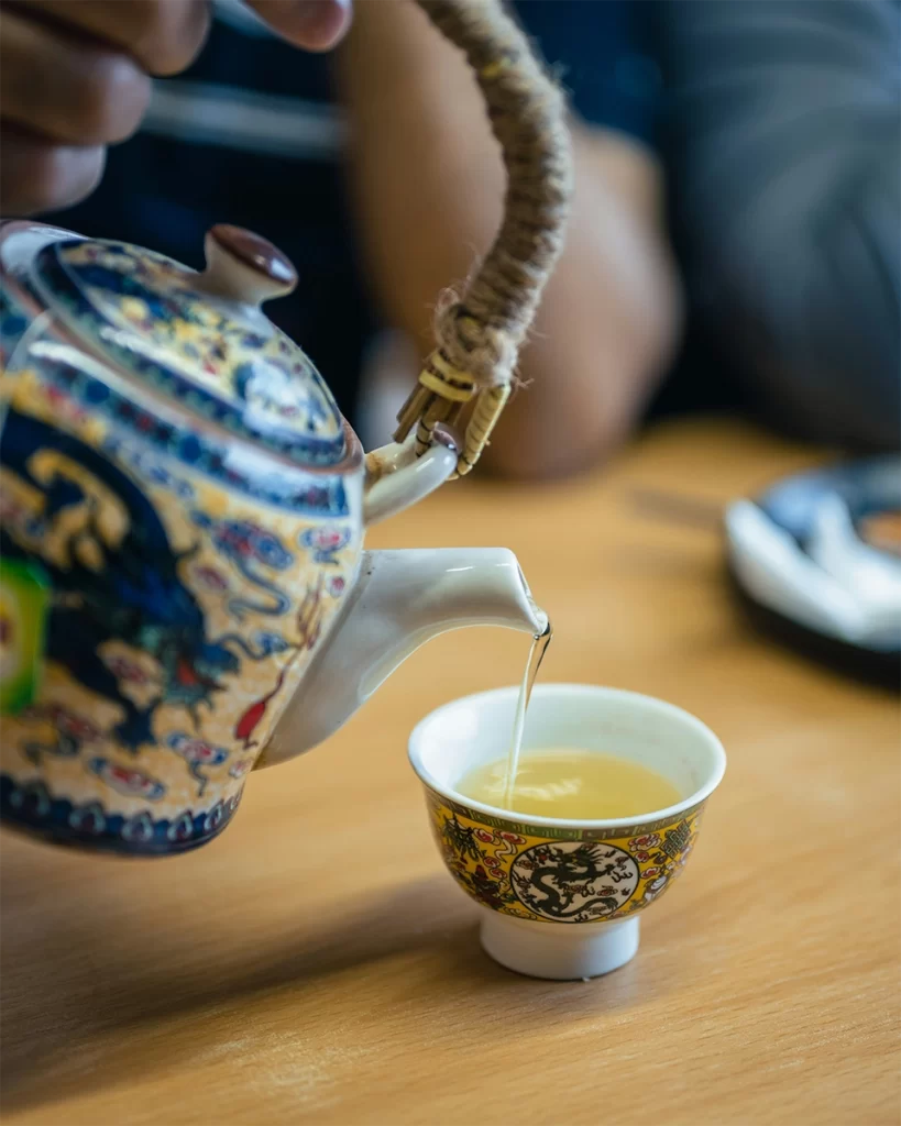A hand pours tea from a beautifully decorated ceramic teapot into a small, intricate teacup with dragon motifs, set on a wooden table in a casual setting. The authentic teaware and setting suggest a local tea experience, offering a quiet alternative to typical tourist trap cafés abroad