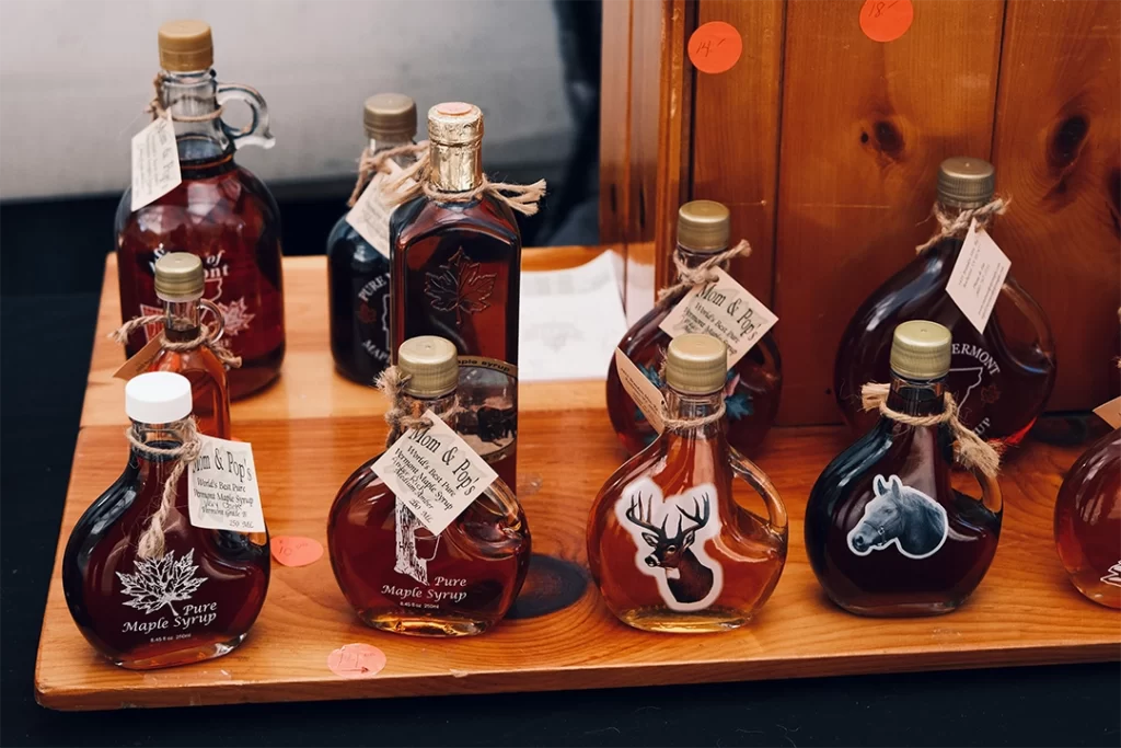 Assorted bottles of pure maple syrup with handcrafted labels and animal decals are displayed on a wooden stand at a market. These natural syrups, like those labeled "Mom & Pop's," highlight maple syrup as a potential best sweetener for gut health thanks to its prebiotic compounds and minimal processing.