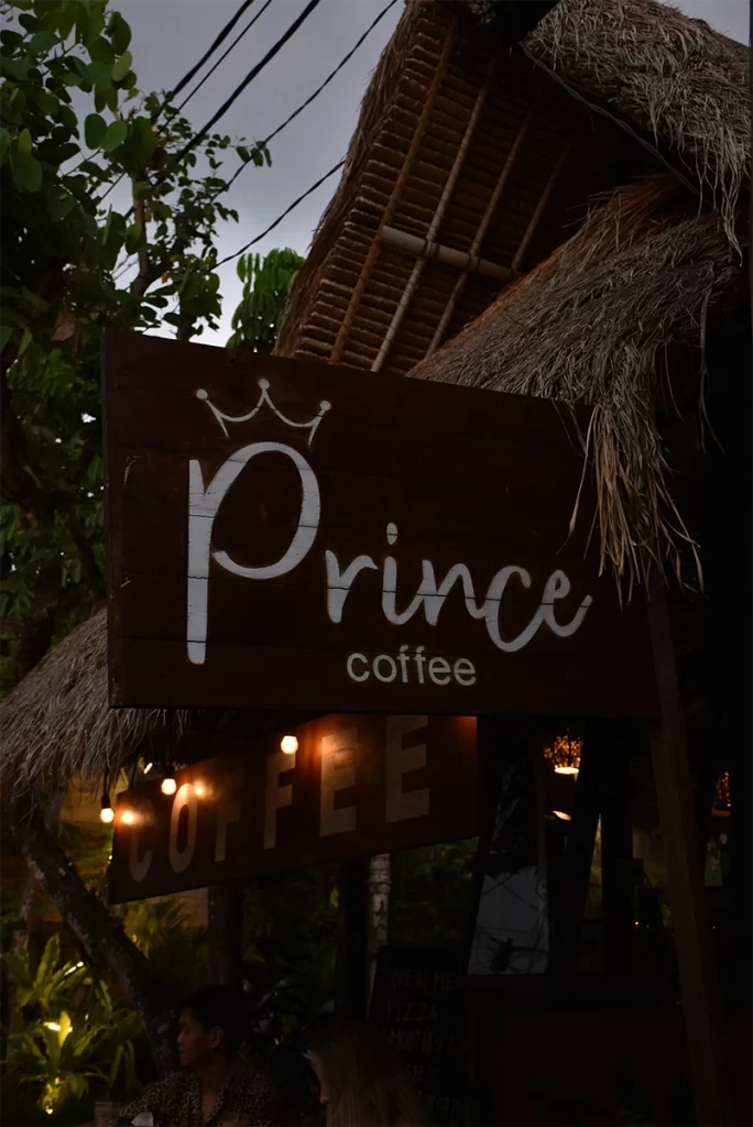 A warmly lit wooden sign reading "Prince Coffee" hangs under a thatched roof with leafy trees and string lights in the background, creating a cozy and inviting vibe. This relaxed, open-air café suggests a senior-friendly atmosphere perfect for enjoying coffee abroad in comfort.